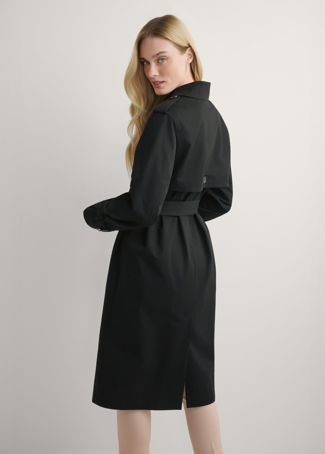 Dlhá čierna dámska bunda v podobe kabáta KURDT-0512-99(W25), pohľad na produkt zozadu na modelke, viditeľná polovica siluety.