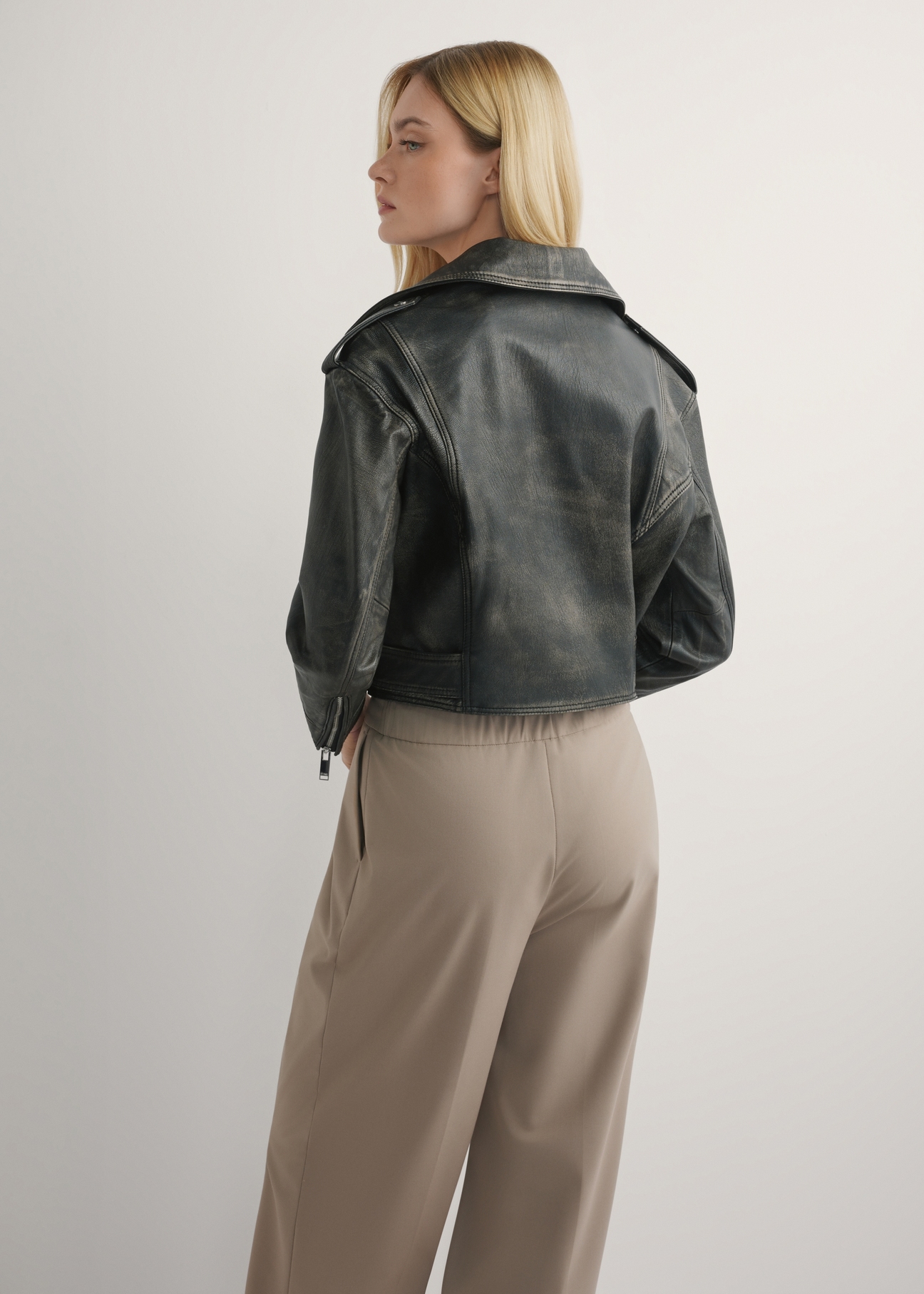 Dámska krátka kožená motorkárska bunda KURDS-0434-99(W26) - pohľad na výrobok zozadu, na modelke, na svetlom pozadí. Viditeľná silueta po kolená.