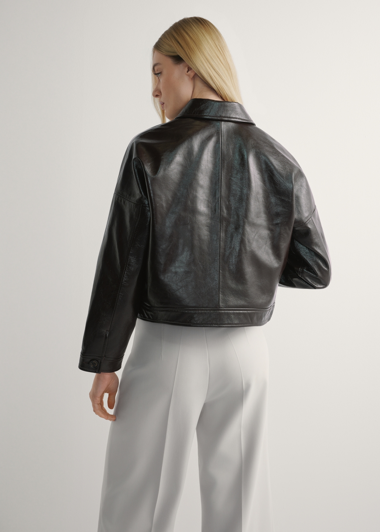 Dámska hnedá kožená krátka bunda KURDS-0593-1I(W26) - pohľad na výrobok zozadu, na modelke, na svetlom pozadí. Viditeľná silueta po kolená.