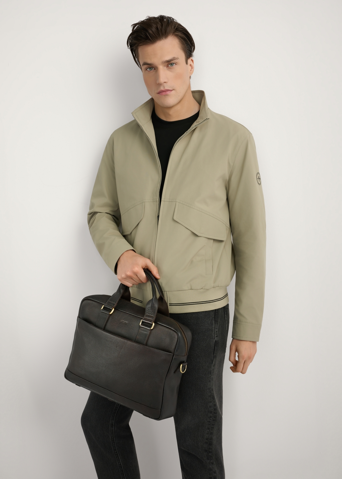 Hnedá pánska kožená aktovka TORMS-0503-1I(W26) - pohľad spredu na produkt, držaný v ruke modelu, na svetlom pozadí, viditeľná silueta po kolená.