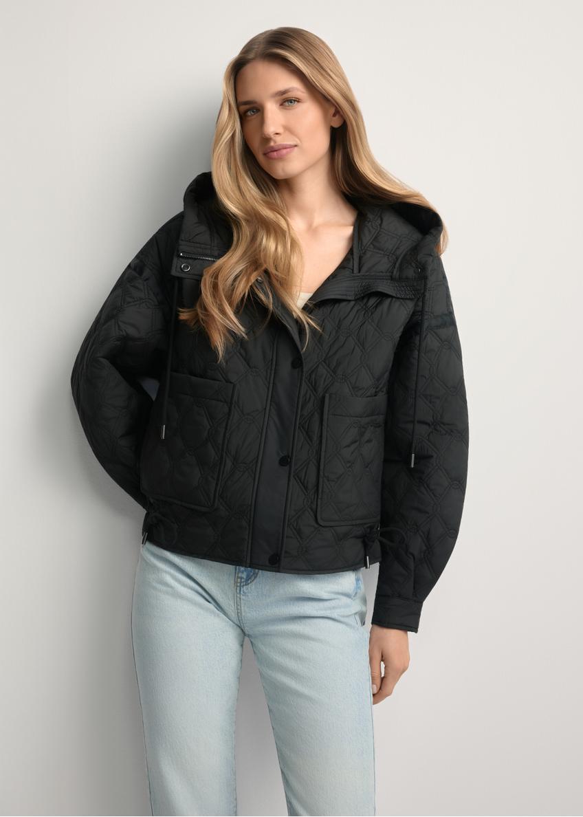 Čierna krátka prešívaná bunda KURDT-0660-99(W26) - pohľad spredu na výrobok, na modelke, na svetlom pozadí. Viditeľná silueta po kolená.